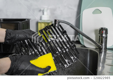 Hands in black gloves diligently cleaning an air fryer grill grate with a yellow sponge and soap in a kitchen sink, for household hygiene Hands in black gloves diligently cleaning an air fryer grill grate with a yellow sponge and soap in a kitchen sink, for household hygiene 128822985