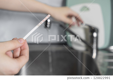 Hand holds a water quality test strip over kitchen sink, checking tap water for purity, ensuring family health and household safety needs Hand holds a water quality test strip over kitchen sink, checking tap water for purity, ensuring family health and household safety needs 128822991