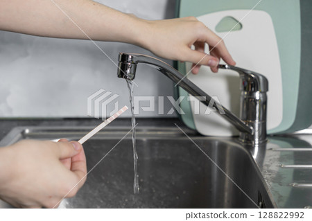 Person's hand holding a water testing strip under running kitchen faucet, analyzing home tap water for drinking safety and quality control Person's hand holding a water testing strip under running kitchen faucet, analyzing home tap water for drinking safety and quality control 128822992