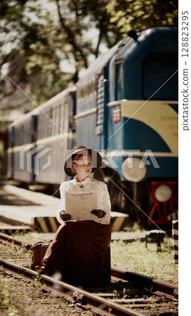 Woman in vintage outfit sitting on tracks reading newspaper beside train 128823295