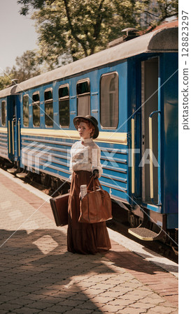 Woman in hat and vintage skirt standing at train platform with luggage and newspaper Woman in hat and vintage skirt standing at train platform with luggage and newspaper 128823297