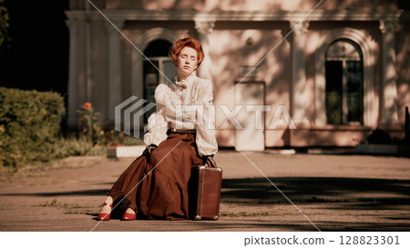 Woman in white blouse and long skirt sitting on pavement with suitcase and red shoes Woman in white blouse and long skirt sitting on pavement with suitcase and red shoes 128823301