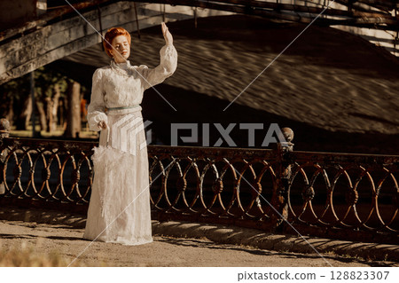 Woman in retro white gown shielding eyes from sun near old bridge railing 128823307