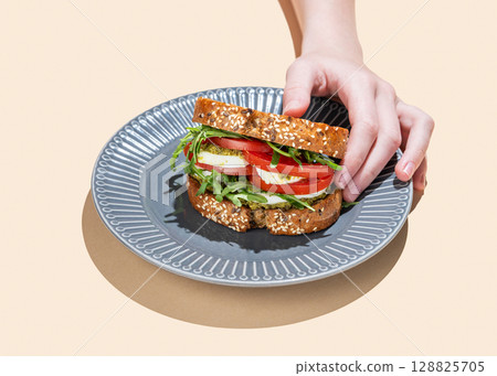 Hand holding sandwich of grain bread with tomatoes, mozzarella, arugula and pesto on a plate Hand holding sandwich of grain bread with tomatoes, mozzarella, arugula and pesto on a plate 128825705