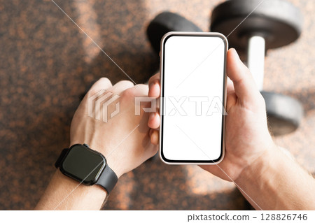 Mans hand holding smartphone with blank screen and wearing smartwatch on wrist, set against background at fitness center. Check fitness progress. Mock up. Mans hand holding smartphone with blank screen and wearing smartwatch on wrist, set against background at fitness center. Check fitness progress. Mock up. 128826746
