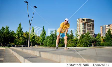 Happy senior man resting with hands on knees after run in modern city park Happy senior man resting with hands on knees after run in modern city park 128828157