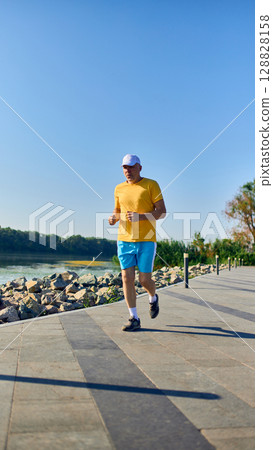 Senior man jogging by riverside in morning sunlight wearing yellow T-shirt Senior man jogging by riverside in morning sunlight wearing yellow T-shirt 128828158