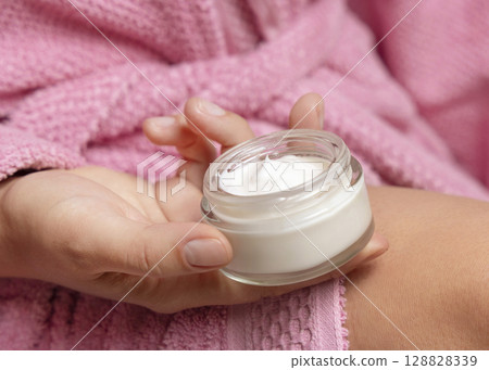 Closeup of young woman in pink bathrobe with opened cream jar in hand. Bodycare concept Closeup of young woman in pink bathrobe with opened cream jar in hand. Bodycare concept 128828339