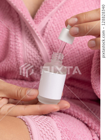 Closeup of woman in pink bathrobe opening cosmetic glass dropper bottle, cosmetic mockup Closeup of woman in pink bathrobe opening cosmetic glass dropper bottle, cosmetic mockup 128828340