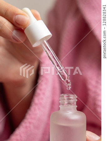 Closeup of woman in pink bathrobe opening cosmetic glass dropper bottle and holding pipette Closeup of woman in pink bathrobe opening cosmetic glass dropper bottle and holding pipette 128828341
