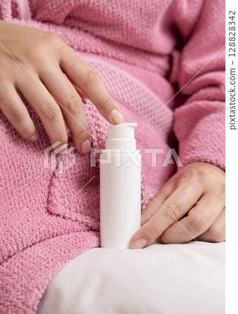 Closeup of young woman in pink bathrobe holding white one pump cosmetic bottle, girly mockup Closeup of young woman in pink bathrobe holding white one pump cosmetic bottle, girly mockup 128828342