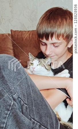 Teenage boy and a cat. Handsome guy hugs his cat while sitting on the couch. Vertical Teenage boy and a cat. Handsome guy hugs his cat while sitting on the couch. Vertical 128828936