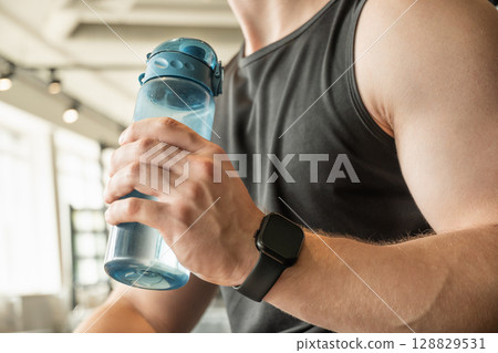 Water bottle in mans hand at gym. Moment of relaxation and refreshment after workout. Close up. 128829531