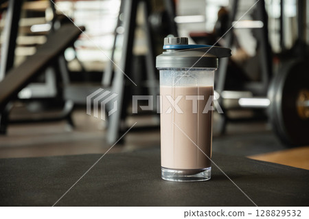Protein shake bottle at gym. Fitness enthusiasm and post-workout routine. Healthy lifestyle and recovery after training. Close up. Protein shake bottle at gym. Fitness enthusiasm and post-workout routine. Healthy lifestyle and recovery after training. Close up. 128829532