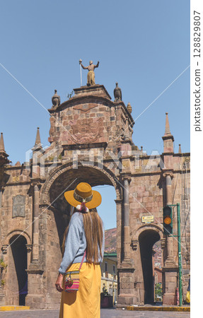 Young woman traveler in brown hat walks through of the Santa Clara Arch in San Francisco square. Cusco, Peru. 128829807