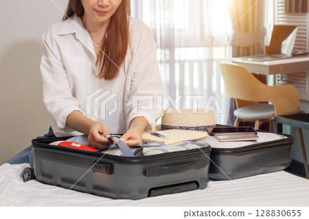 happy Asian woman packing suitcase on bed in bedroom, preparing for summer holidays abroad trip with passport, hat, travel medicine first aid kit, cloth, phone, notebook for checklist 128830655
