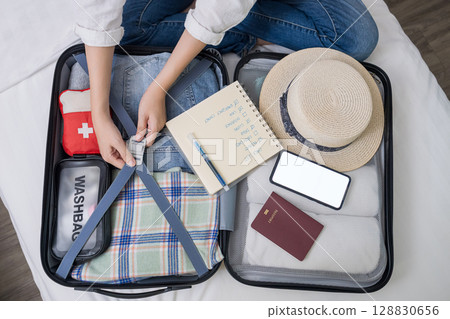 top view of woman hand packing suitcase for trip, preparing for summer holidays abroad with travel medicine first aid kit, cloth, accessory, phone mockup on open luggage, notebook for checklist item 128830656
