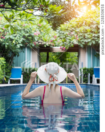 back view of woman in pink swimsuit with straw hat relaxing in swimming pool at hotel, enjoy summer holiday and vacation at tropical country, sunbathing wellness lifestyle concept. 128830658