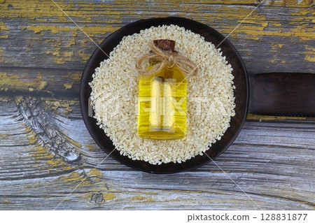Glass bottle of sesame oil and raw sesame seeds in wooden spoon or bowl on rustic table. Uncooked sesame background concept with copy space Glass bottle of sesame oil and raw sesame seeds in wooden spoon or bowl on rustic table. Uncooked sesame background concept with copy space 128831877