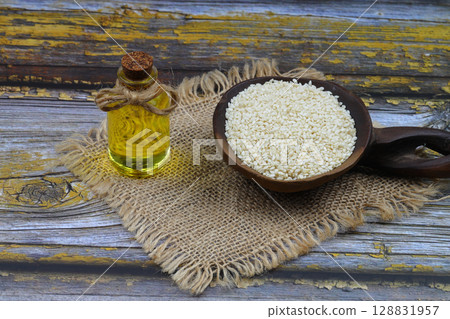 Glass bottle of sesame oil and raw sesame seeds in wooden spoon or bowl on rustic table. Uncooked sesame background concept with copy space Glass bottle of sesame oil and raw sesame seeds in wooden spoon or bowl on rustic table. Uncooked sesame background concept with copy space 128831957