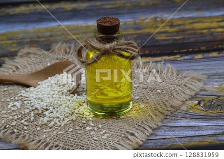 Glass bottle of sesame oil and raw sesame seeds in wooden spoon or bowl on rustic table. Uncooked sesame background concept with copy space 128831959