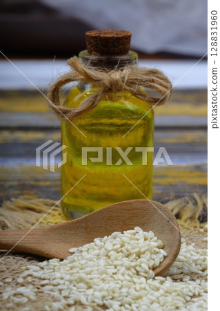 Glass bottle of sesame oil and raw sesame seeds in wooden spoon or bowl on rustic table. Uncooked sesame background concept with copy space Glass bottle of sesame oil and raw sesame seeds in wooden spoon or bowl on rustic table. Uncooked sesame background concept with copy space 128831960