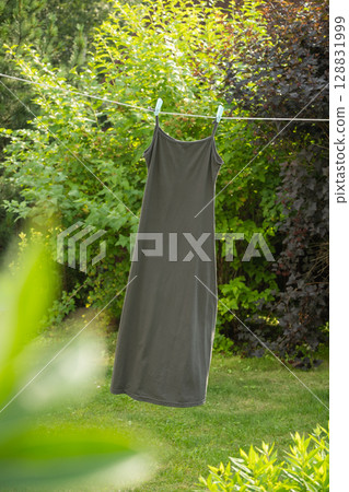 Gray sleeveless shirt hanging on clothesline with clothespins in outdoor garden. Natural environment creates serene and refreshing atmosphere for air drying. Gray sleeveless shirt hanging on clothesline with clothespins in outdoor garden. Natural environment creates serene and refreshing atmosphere for air drying. 128831999