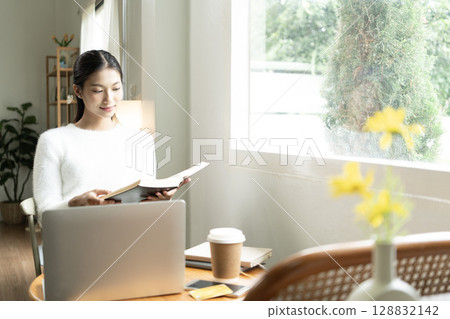 Young Asian woman enjoying a peaceful moment reading a book near window. Calm lifestyle, self-learning, and mindfulness at home. 128832142