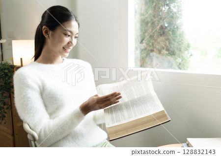 Young Asian woman enjoying a peaceful moment reading a book near window. Calm lifestyle, self-learning, and mindfulness at home. 128832143