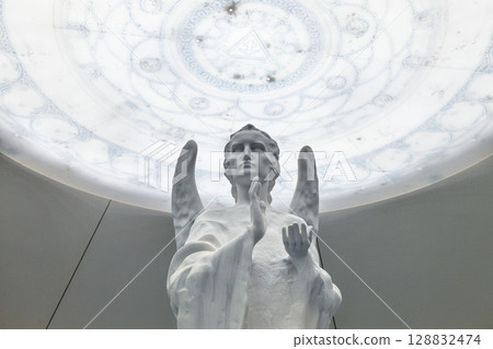 Angelic white statue praying beneath ornate luminous dome in modern sacred interior 128832474
