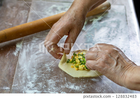 Process of preparing pelmeni with vegetable filling of onions and eggs on floured surface. Close-up of an elderly woman's hands 128832483