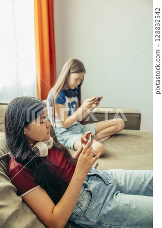 two girls relaxing on sofa using smartphone, headphones, casual home setting bright curtains and natural light window modern lifestyle, technology education entertainment 128832542