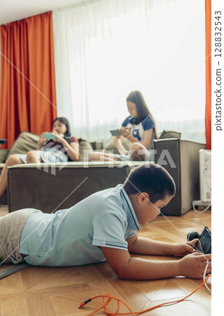 boy lying on wooden floor playing with tablet while two girls relax on sofa using smartphones with orange curtains, daylight, technology education leisure, vertical boy lying on wooden floor playing with tablet while two girls relax on sofa using smartphones with orange curtains, daylight, technology education leisure, vertical 128832543