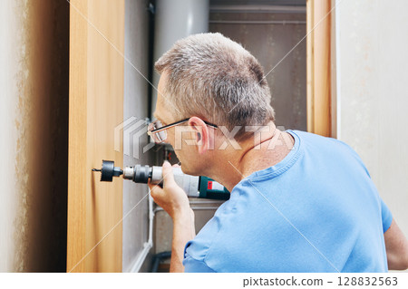 Drilling hole in wooden door using hole saw mounted in drill chuck for installation of latch mechanism during door hardware fitting. Drilling hole in wooden door using hole saw mounted in drill chuck for installation of latch mechanism during door hardware fitting. 128832563