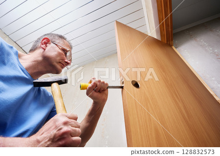 Creating precise mortise with chisel and hammer, installer shapes cavity inside door edge to fit lock mechanism as part of interior door renovation. 128832573