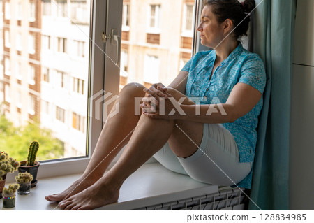 Woman sitting on windowsill looking out at city view 128834985