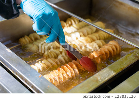 Chef frying tornado potatoes on a stick using tongs in deep fryer Chef frying tornado potatoes on a stick using tongs in deep fryer 128835126