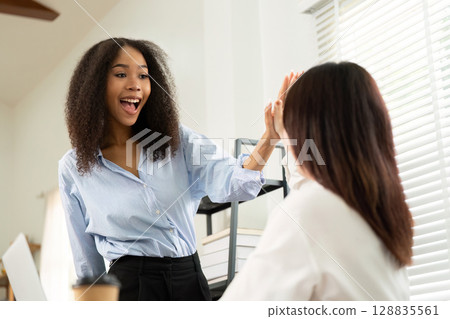 Joyful Interaction. Two women sharing a high-five in a bright office. 128835561