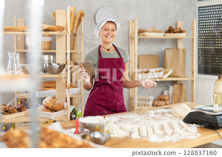 Female baker cutting raw dough with knife on kitchen table in private bakery 128837160