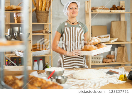 Young woman baker are standing near product range in bakery kitchen with basket of bread. Young woman baker are standing near product range in bakery kitchen with basket of bread. 128837751