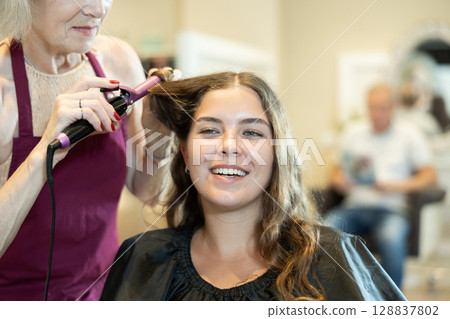 In beauty salon, cropped elderly female hairdresser makes styling curls for young girl client 128837802