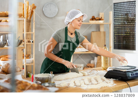 Female baker cutting raw dough with knife and weighs pieces of raw dough on scales for making croissants Female baker cutting raw dough with knife and weighs pieces of raw dough on scales for making croissants 128837809