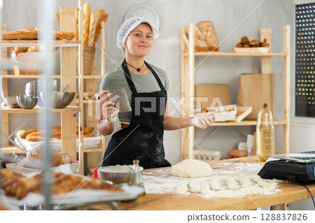 Young baker girl is standing near work table in kitchen, working with dough Young baker girl is standing near work table in kitchen, working with dough 128837826