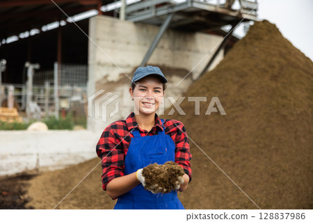Girl worker in blue jumpsuit stands next to pile of manure. 128837986