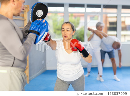 Old woman training boxing kicks on punch mitts held by instructor Old woman training boxing kicks on punch mitts held by instructor 128837999