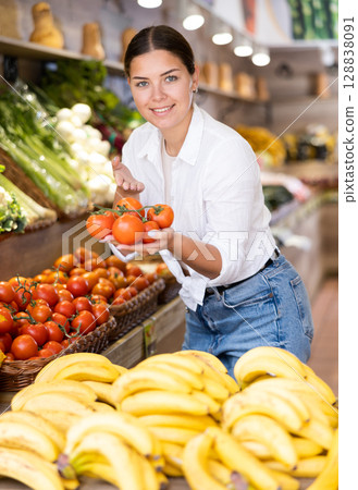 Smiling young woman purchaser holding tomato in grocery store 128838091