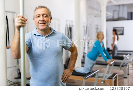 Portrait of smiling elderly male pensioner in activewear posing in modern Pilates fitness studio Portrait of smiling elderly male pensioner in activewear posing in modern Pilates fitness studio 128838107