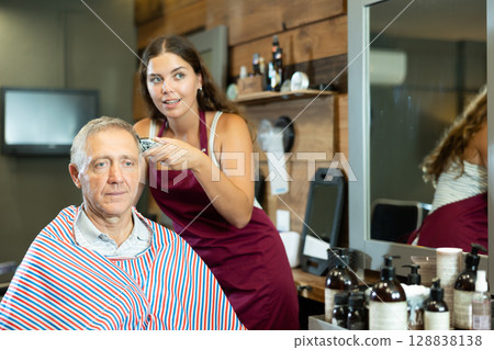 Young female barber making haircut to elderly client in barbershop Young female barber making haircut to elderly client in barbershop 128838138