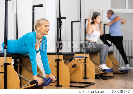 Sporty mature female doing stretching exercises on pilates reformer Sporty mature female doing stretching exercises on pilates reformer 128838187