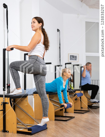 Positive young female performing standing Pilates exercise for legs and core using platform on Wanda chair in gym 128838207